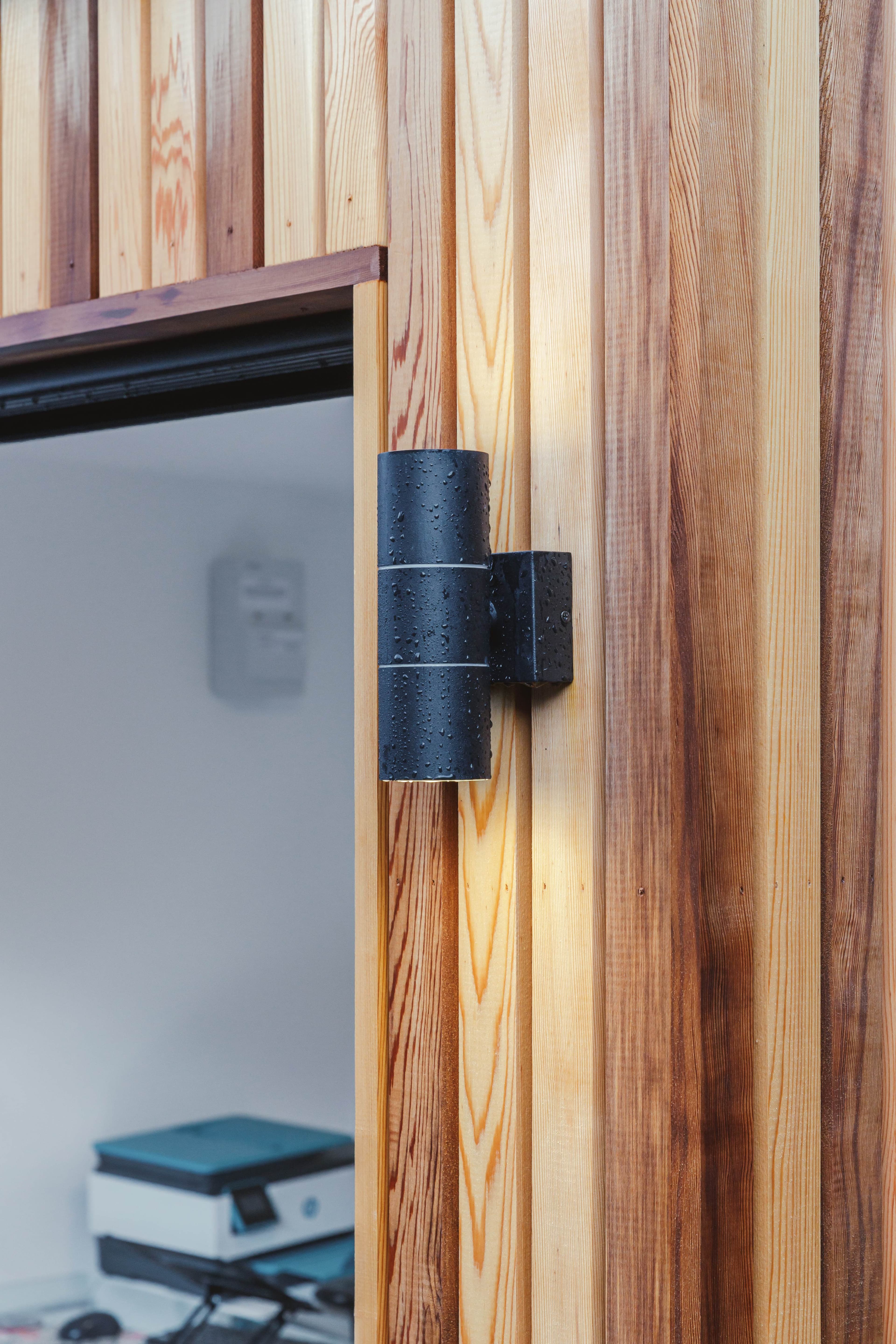 Close-up of a black up-and-down wall light on cedar cladding beside the open doorway of a Bedfordshire garden room