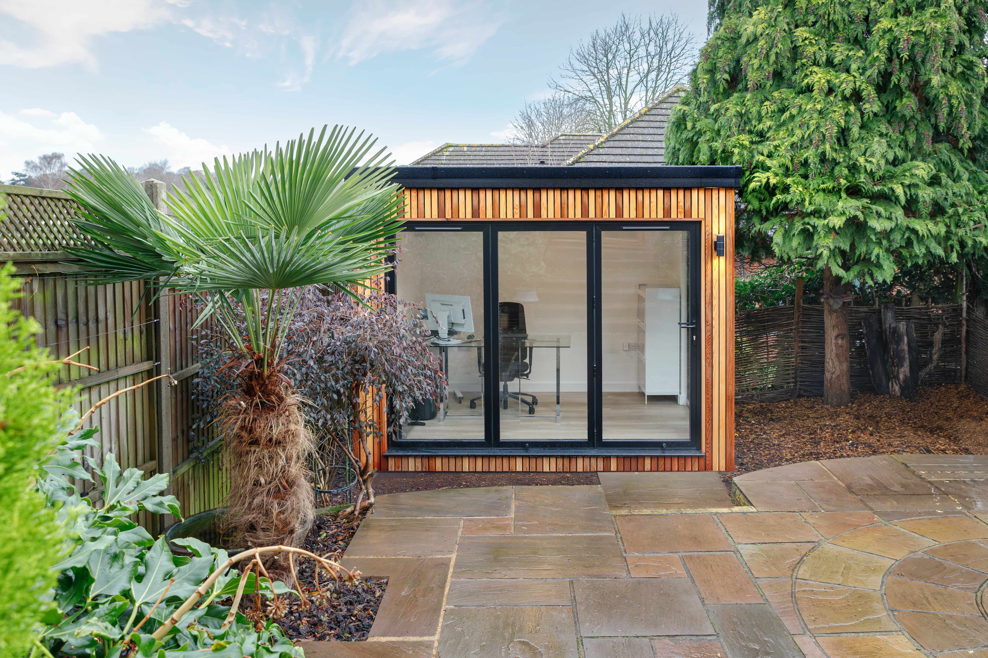 Cedar-clad garden office in Bedfordshire with black bifold doors, a desk and office chair inside, on a sandstone patio beside a palm