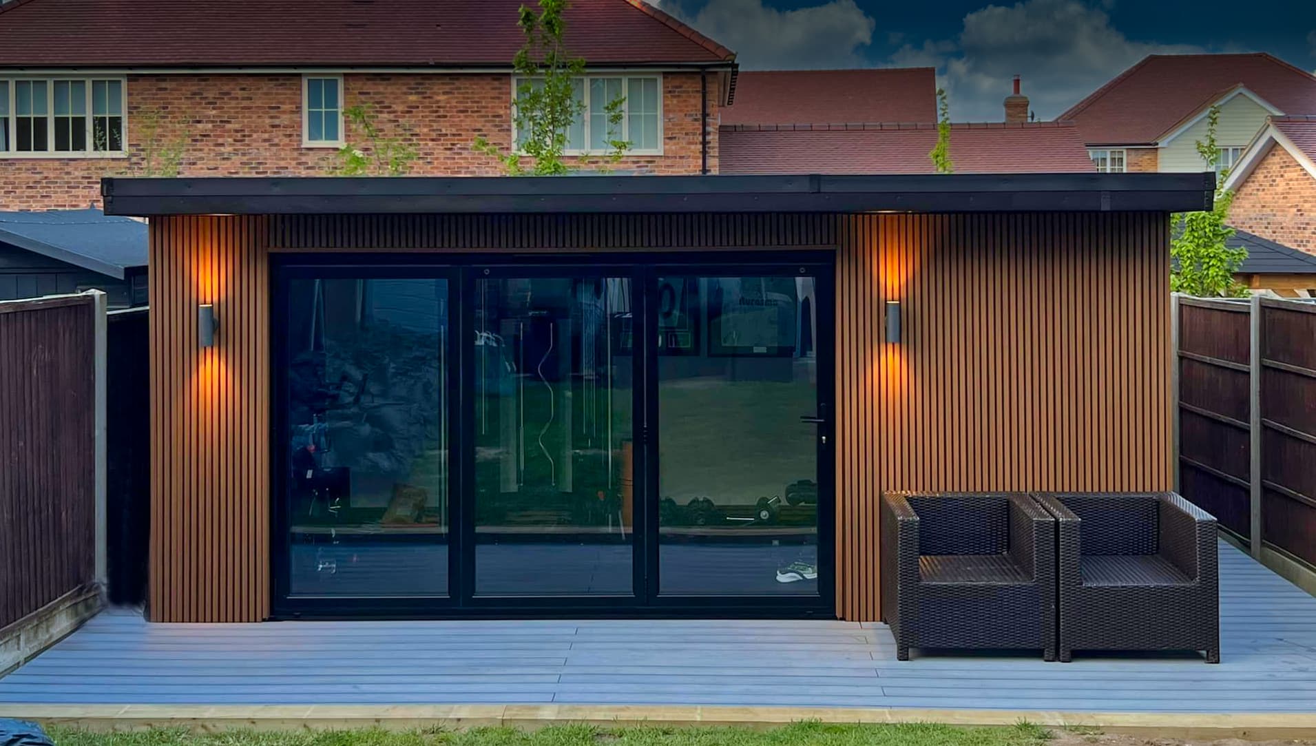 Cedar slatted garden room in Bedfordshire lit warmly at dusk with rattan seating on the deck