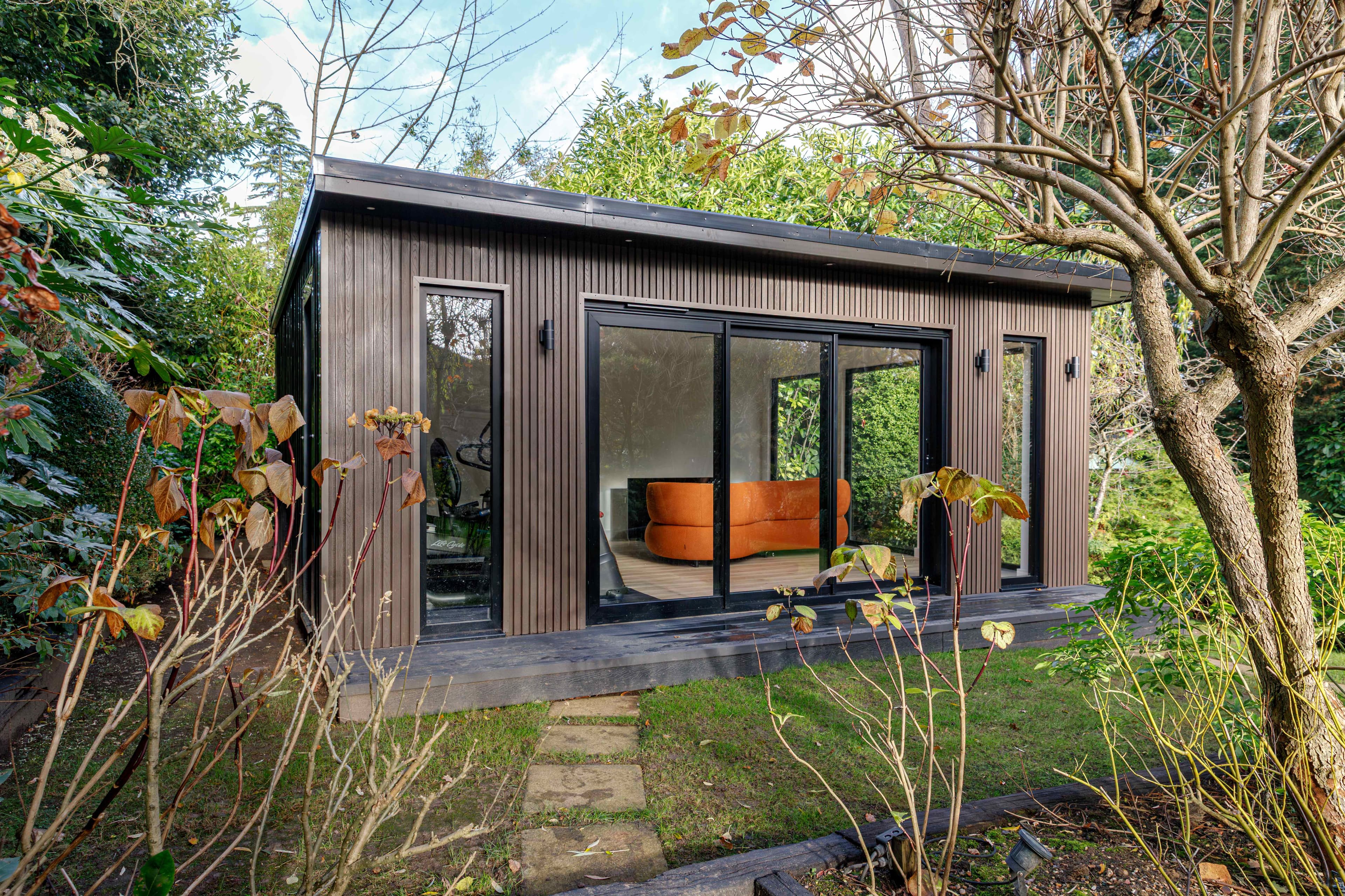 Composite-clad garden room in Bedfordshire with large sliding doors and a curved orange sofa inside, framed by autumn planting
