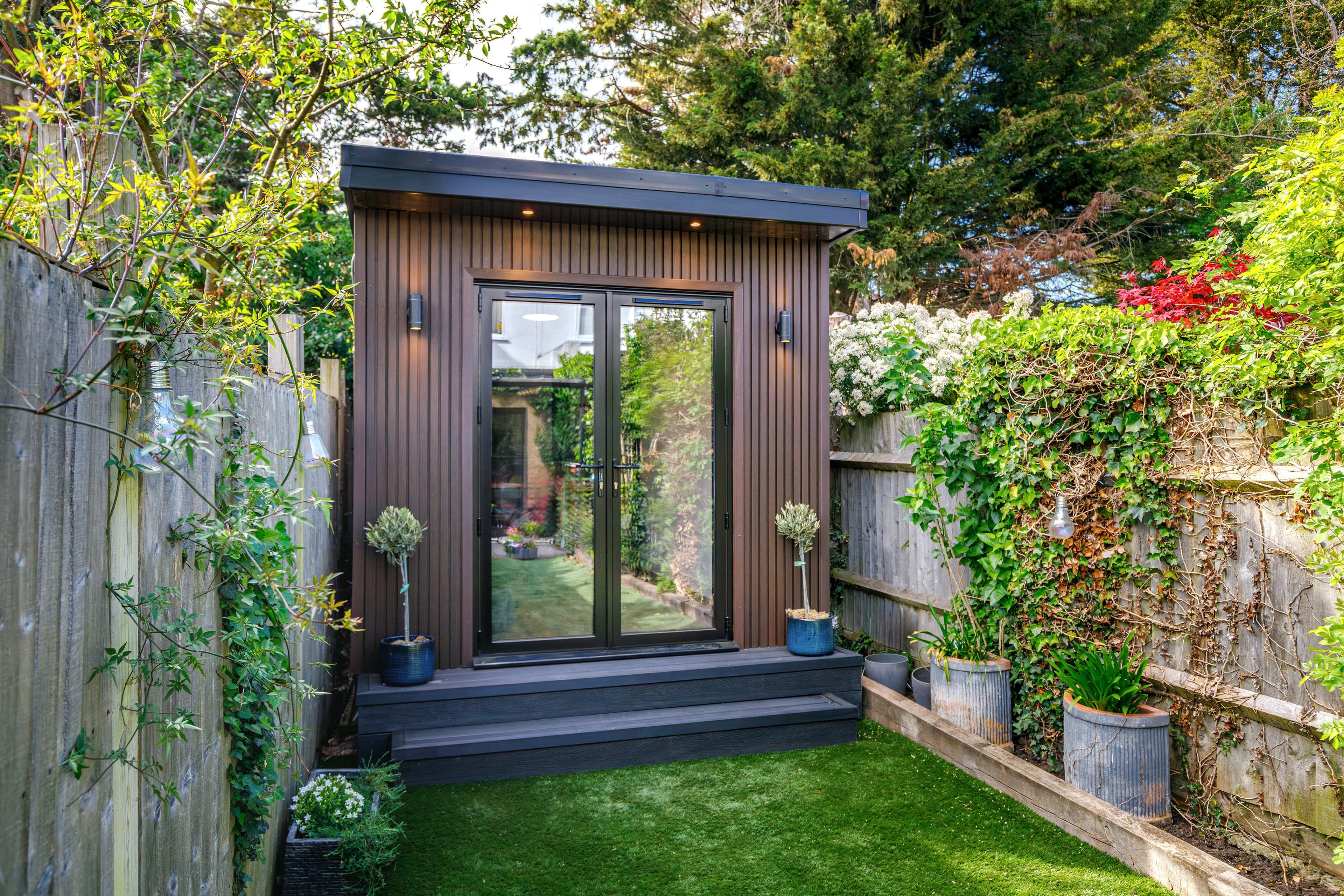 Dark timber-clad garden room in Bedfordshire with black French doors, flanked by potted olive trees on a small lawn