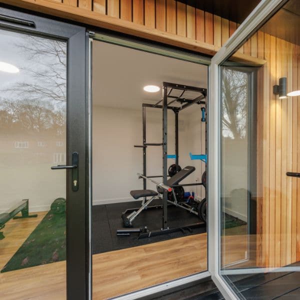 View through open doors into a Bedfordshire garden gym fitted with a power rack, weight bench and cardio equipment