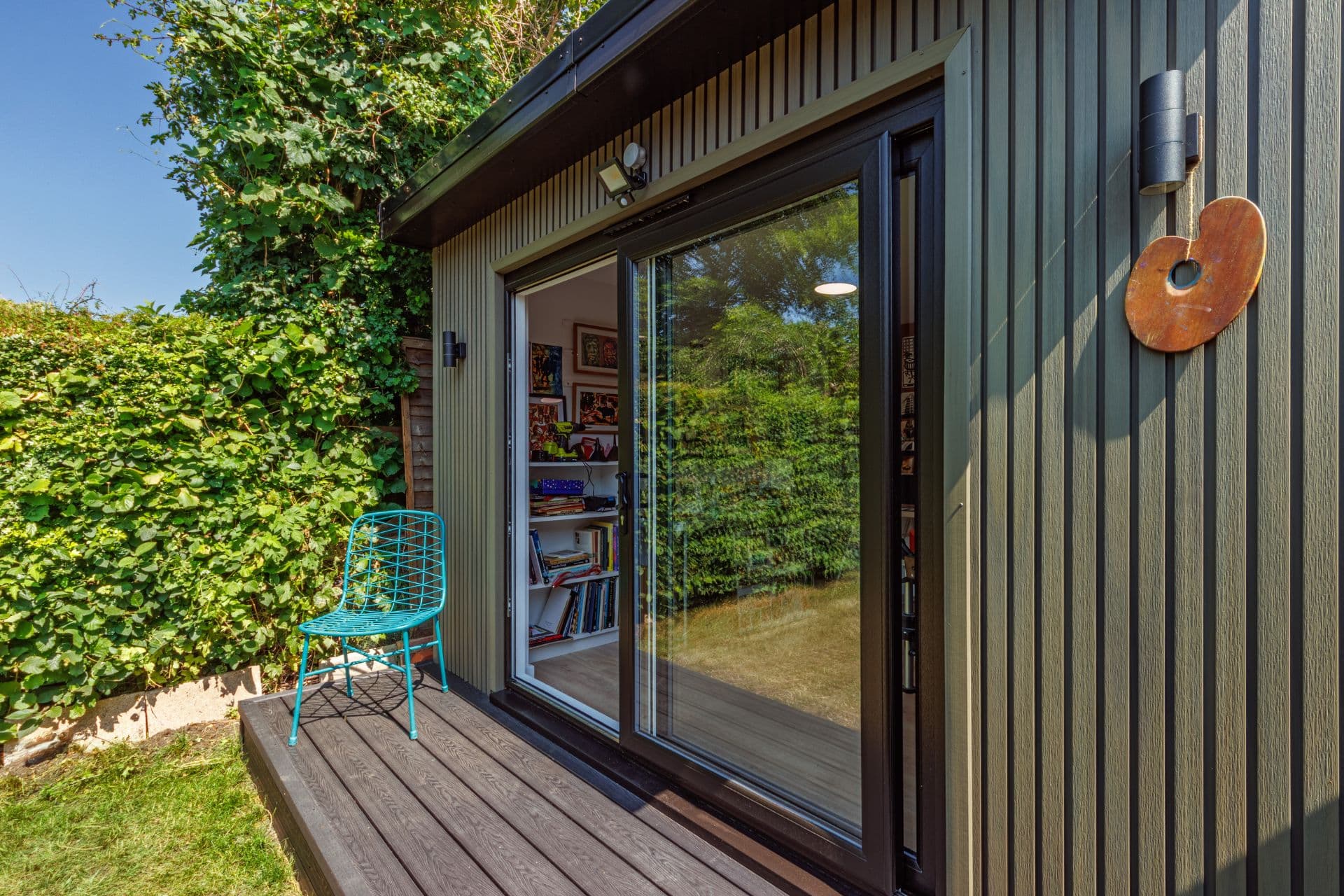 Green composite-clad garden art studio in Bedfordshire with a sliding door open to a book-lined interior