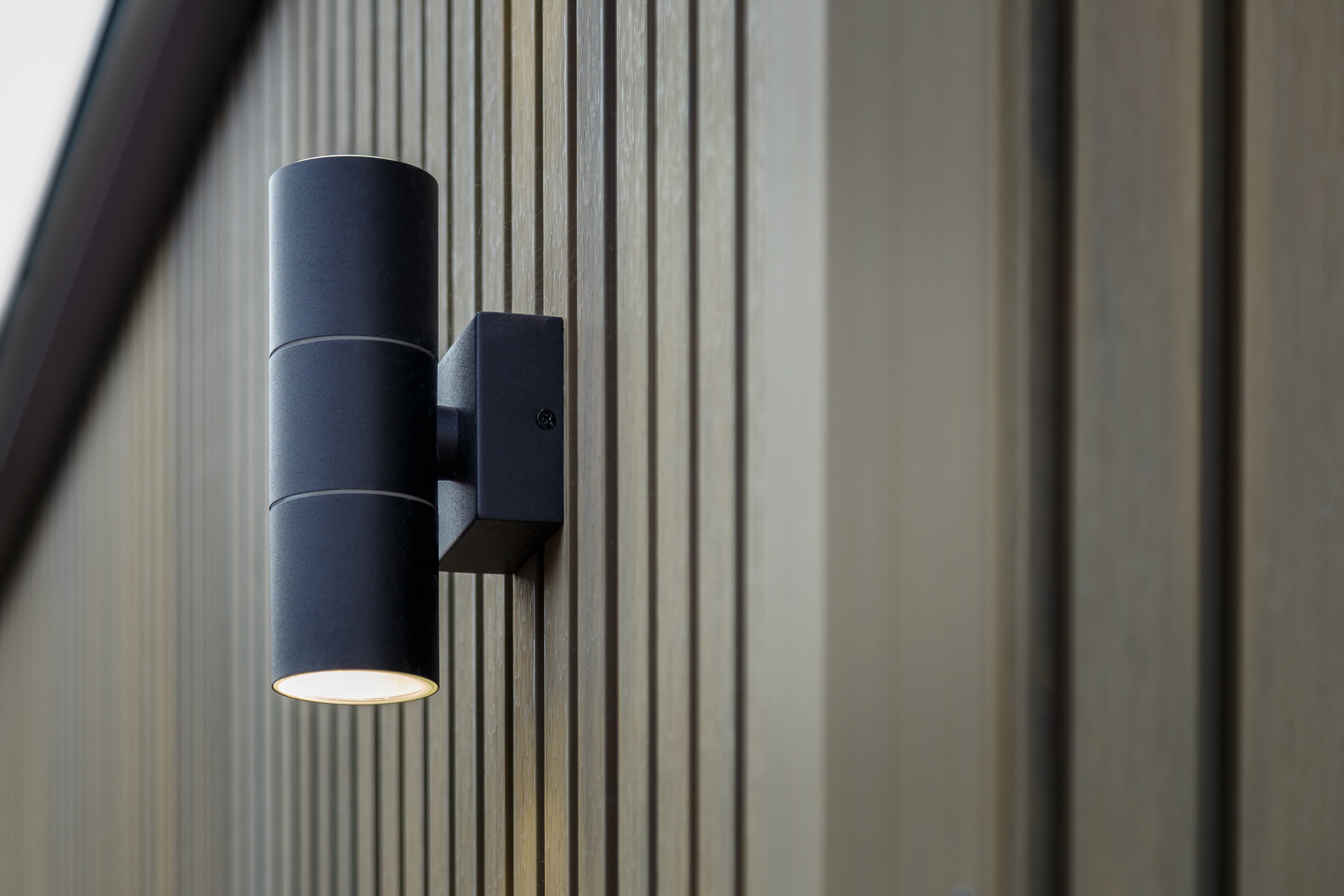 Close-up of a black cylindrical up-and-down wall light mounted on greige slatted composite cladding of a Bedfordshire garden room