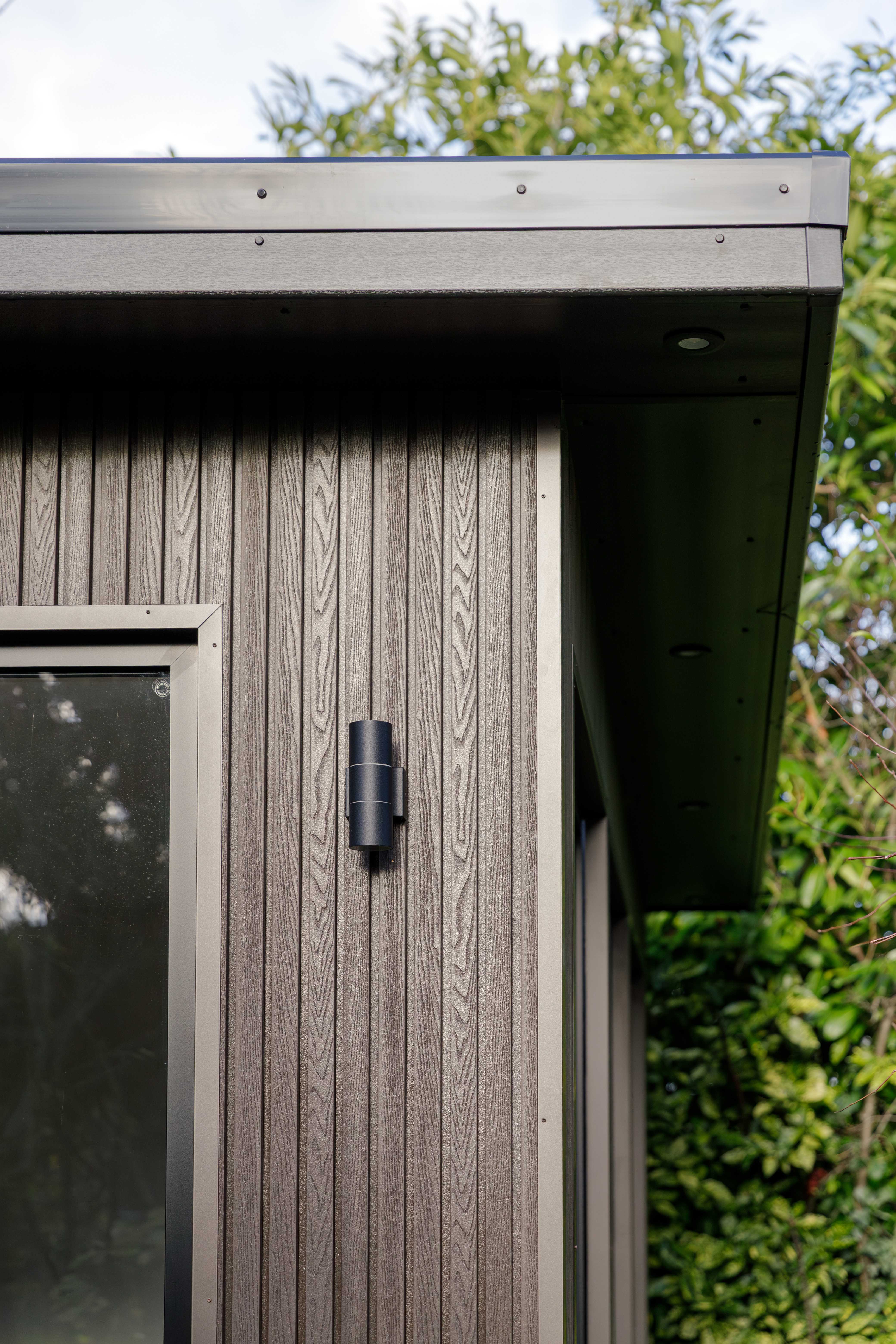 Corner detail of grey wood-grain composite cladding on a Bedfordshire garden room with a black cylindrical wall light and dark roof trim