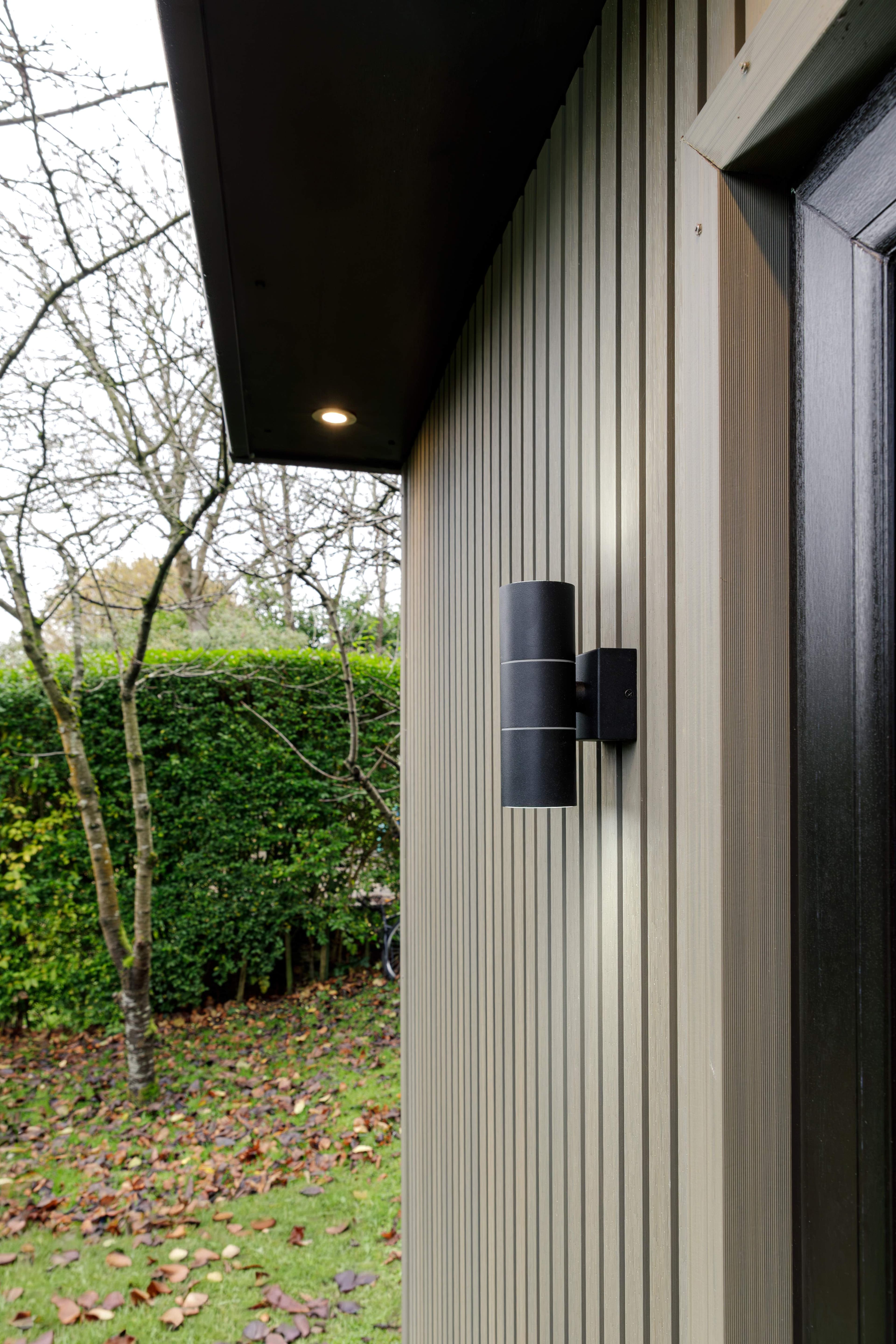 Black up-and-down wall light on pale grey slatted cladding beneath the roof overhang of a Bedfordshire garden room