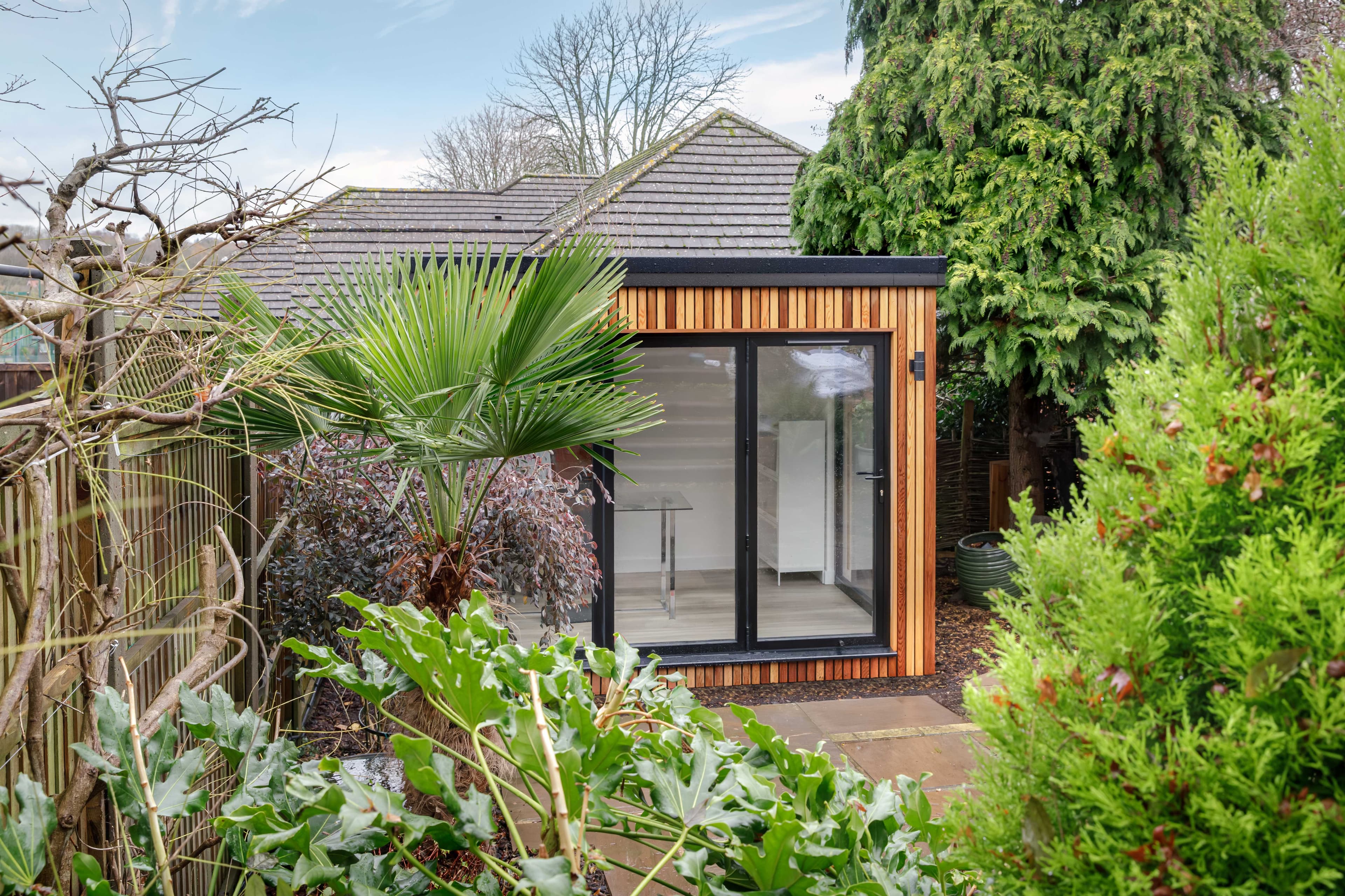 Cedar-clad garden room in Bedfordshire with corner glazing and sliding doors, framed by lush palm and shrub planting
