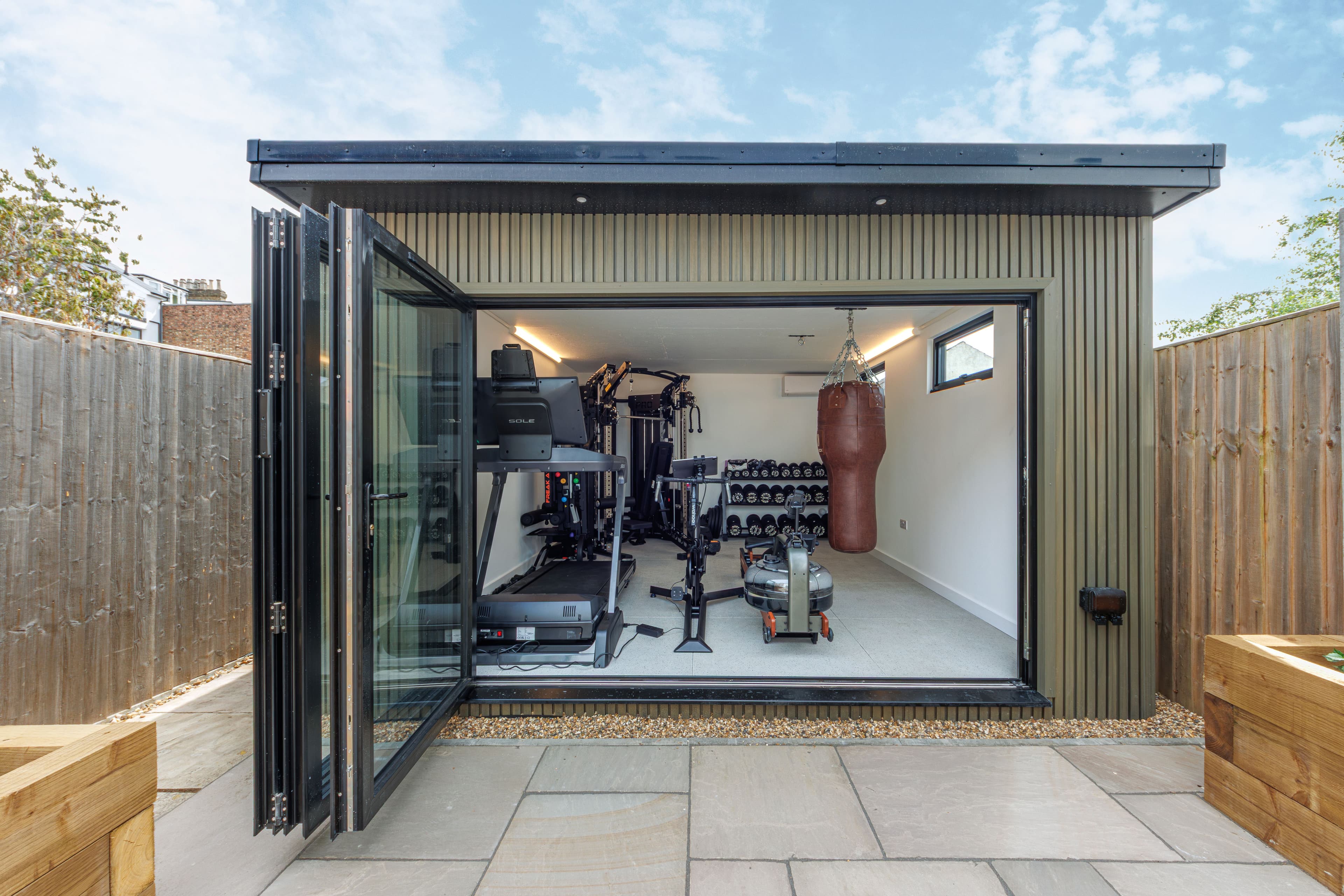 Garden gym in Flitwick — olive corrugated-clad building with bifold doors open onto a paved patio, fitted with a treadmill, cable machine, dumbbells and a punchbag
