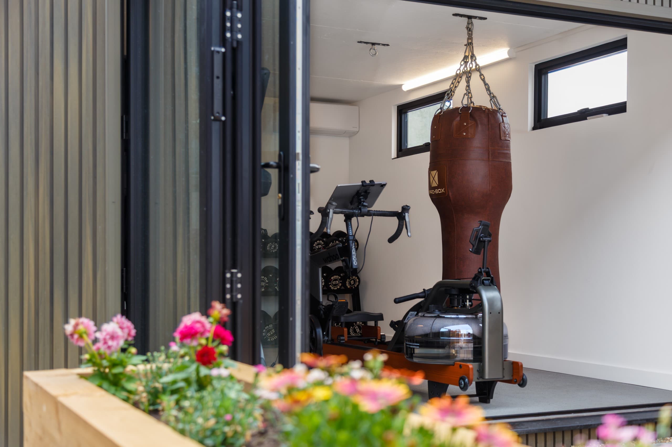 Olive-clad garden gym in Bedfordshire seen through open bifold doors, with a hanging punchbag, water rower and dumbbell rack beside flower planters