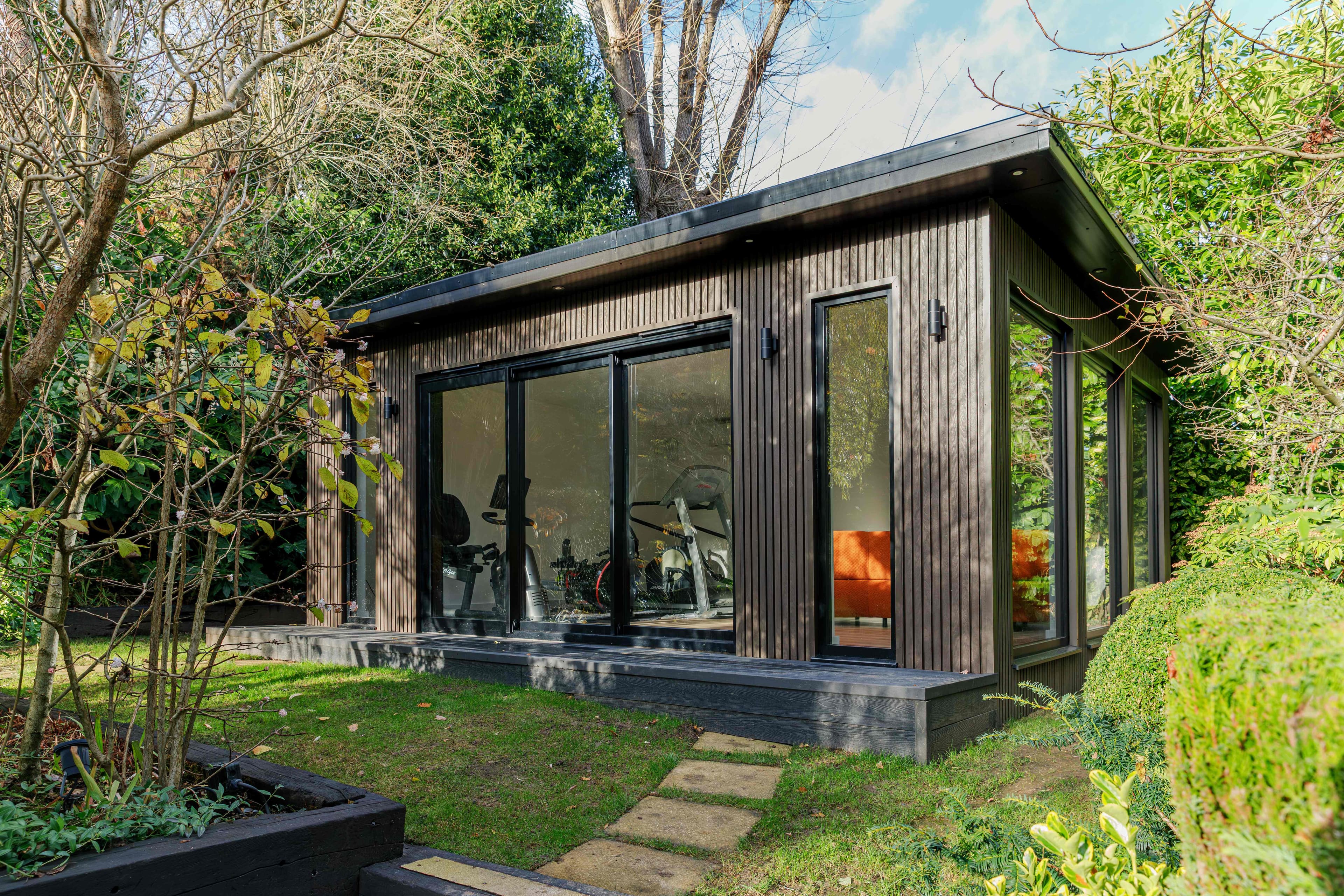 Insulated garden room in Bedford — dark timber slatted cladding with corner glazing and sliding glass doors, set on a deck in a leafy garden